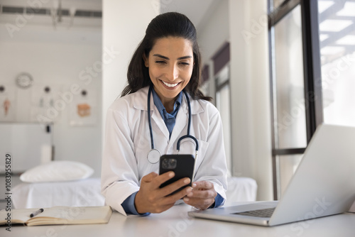Wallpaper Mural Pretty Latina female doctor in white medical coat sit at desk in hospital look at cellphone screen browse wireless internet, consult clinic patient online via smartphone telehealth mobile application Torontodigital.ca