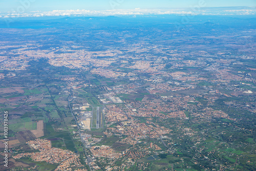 Aerial photo shows a city with dense buildings, green spaces, and a central airport. Surrounding areas include residential zones, fields, and forests. Lazio aerial view in Italy