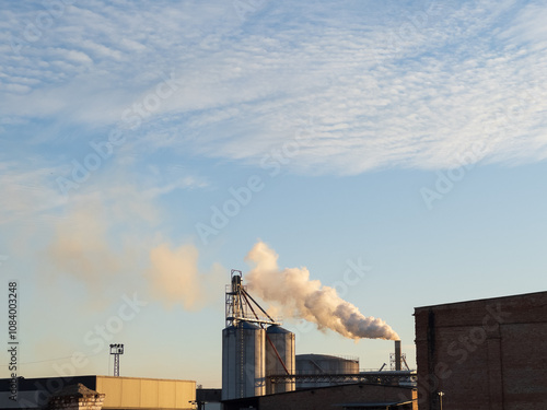 Against a backdrop of a stunning blue sky, towering smokestacks release puffs of smoke as the sun sets, casting a warm glow over the industrial landscape and evoking a sense of transformation photo