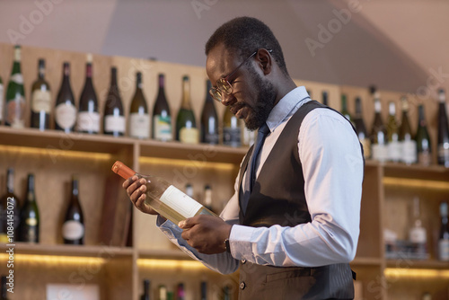 Low angle shot of African American highly skilled sommelier reading label of dry white wine while standing in wine cellar before degustation photo