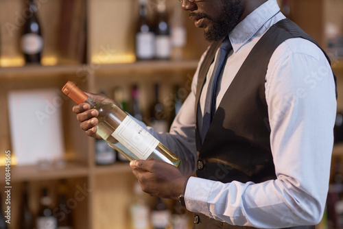 Unrecognizable African American sommelier holding bottle of white while reading label in wine cellar photo