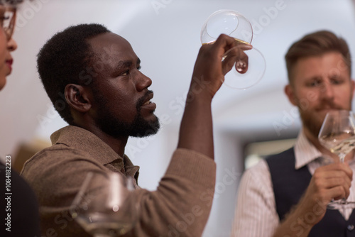 Low angle shot of African American highly skilled sommelier looking at polished crystal glass with white whine while estimating its color photo