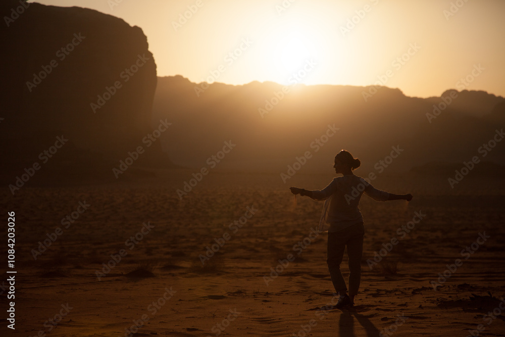 Fototapeta premium silhouette of a young beautiful girl scattering sand from her hands in the wind while being in desert during the sunset