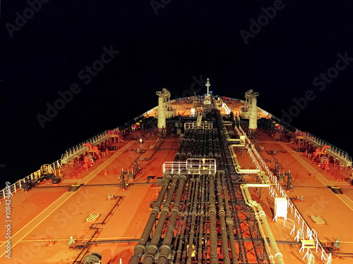 the illuminated deck of an oil tanker at night, highlighting its robust structure and extensive pipeline systems running along the ship's length. The scene is framed by the darkness of the ocean.