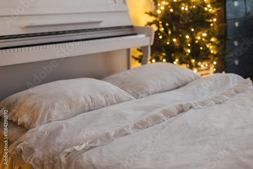 A comfortable bed featuring crisp white sheets and fluffy pillows is positioned next to a beautiful piano, accompanied by a festive Christmas tree