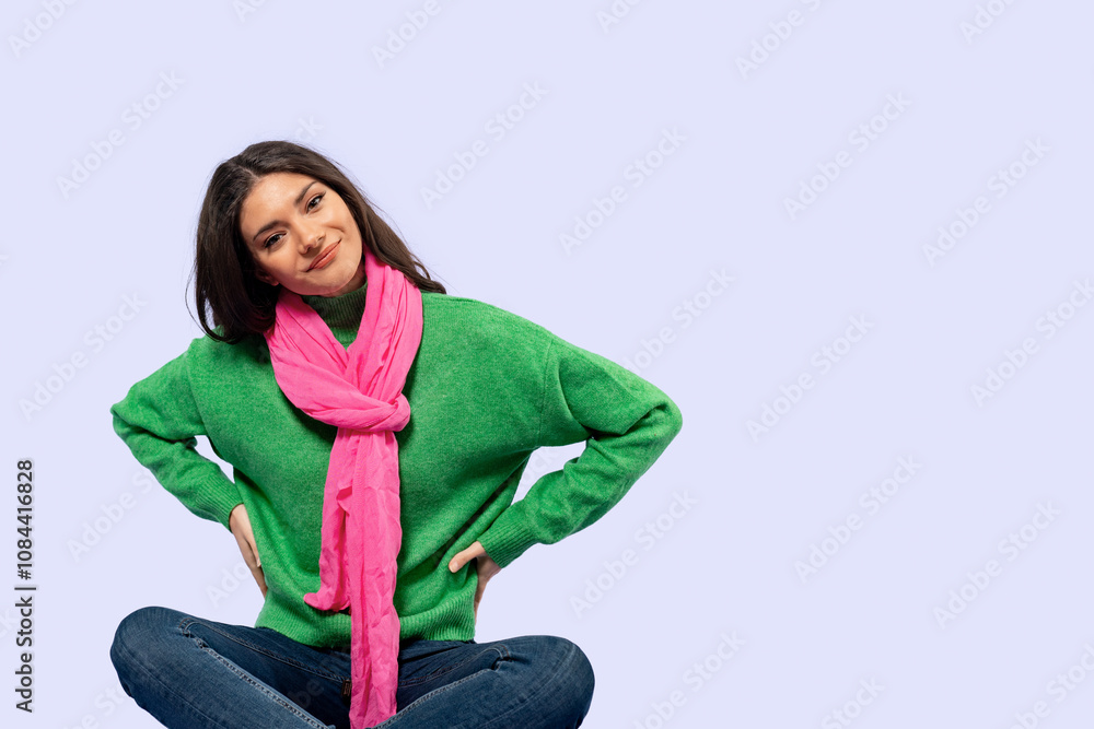 Young woman in a green sweater and pink scarf sitting cross-legged against a pastel background while smiling Young woman in a green sweater and pink scarf sitting cross-legged against a pastel background while smiling