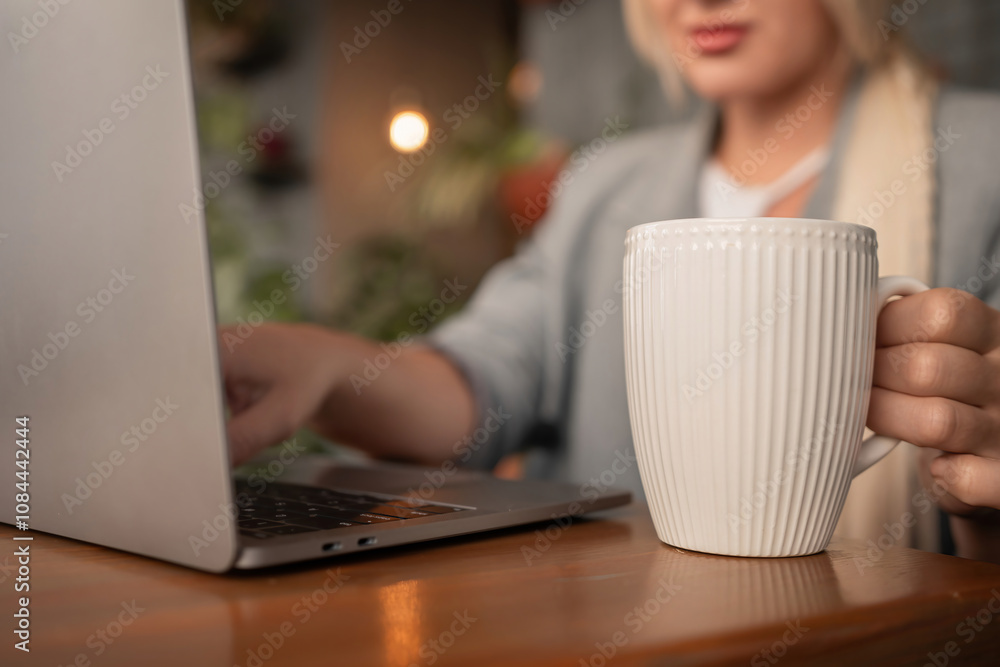A woman is sitting at a desk with a laptop and a white coffee mug. She is typing on the laptop and holding the coffee mug in her hand. Concept of productivity and focus. A woman is sitting at a desk with a laptop and a white coffee mug. She is typing on the laptop and holding the coffee mug in her hand. Concept of productivity and focus.