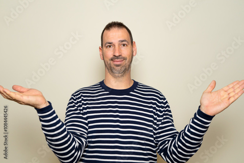 Half length portrait of a bearded hispanic man in his 40s smiling and excited wearing striped sweater and open hands gesture on beige studio background photo