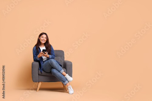 Smiling woman with smartphone sitting in armchair on beige background. Space for text