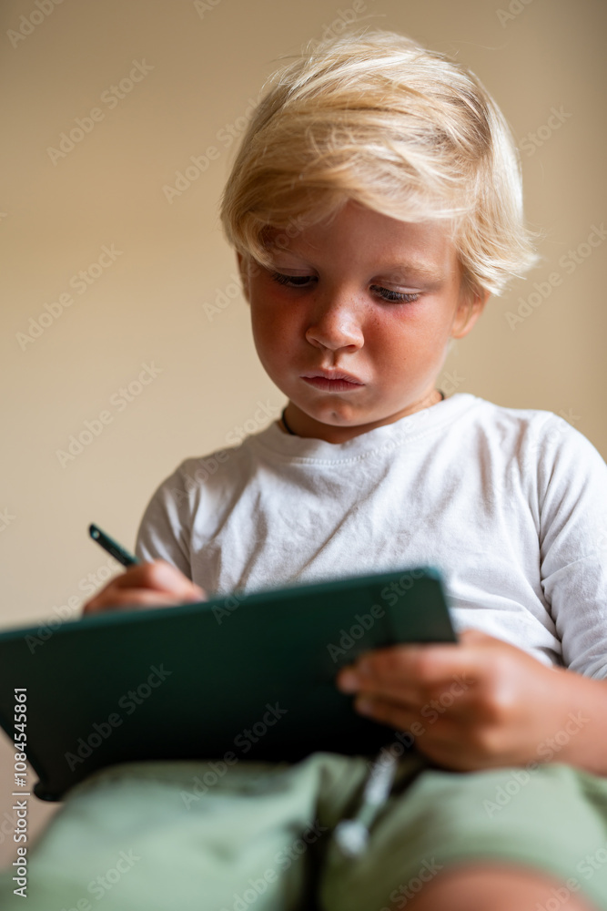 Young Blonde Boy Intently Drawing on a Tablet While Sitting Indoors in a Cozy Living Room Young Blonde Boy Intently Drawing on a Tablet While Sitting Indoors in a Cozy Living Room