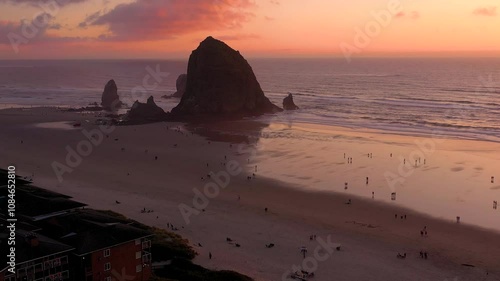 Aerial drone footage flying away and revealing the scenic Haystack Rock in Cannon Beach, Oregon during a vibrant sunset.
