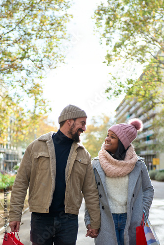 Vertical. Beautiful young couple interracial in love enjoying winter holiday season in the city streets, taking a walk, hugging and holding gift bags while celebrating Christmas outdoors