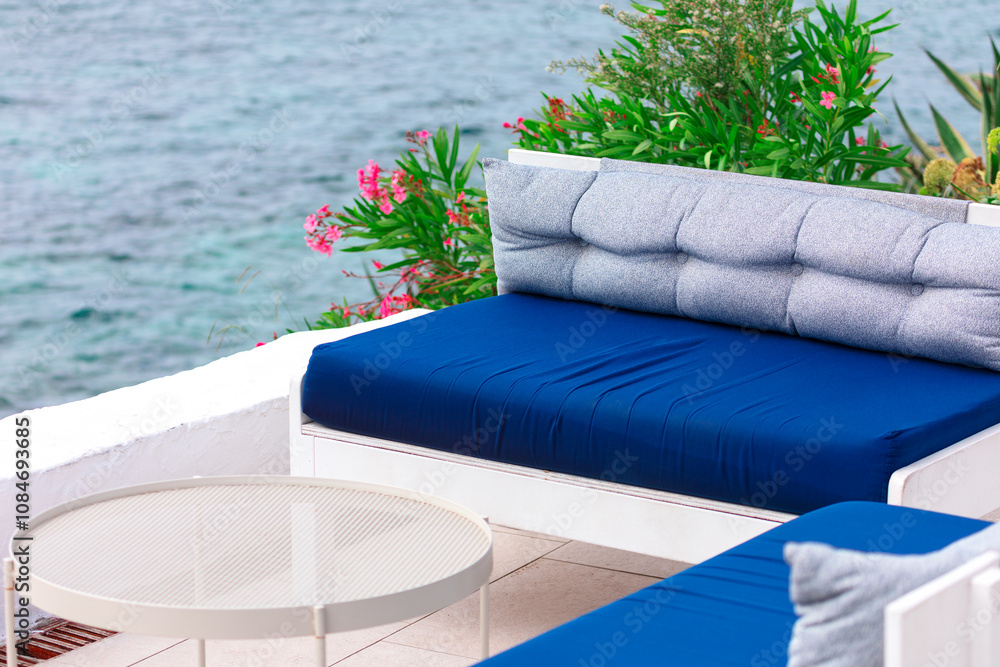 Outdoor seating area overlooks a calm, blue sea. A white-framed couch with blue and gray cushions sits by a round white table, surrounded by green plants with pink flowers, creating a serene, relaxing