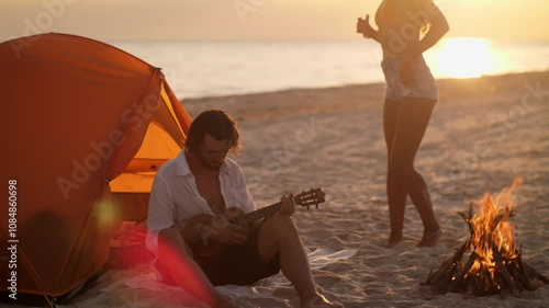 A serene beach scene at sunset with a man playing the ukulele and a woman dancing near a campfire. Captures the essence of carefree summer evenings, perfect for travel and lifestyle concepts.