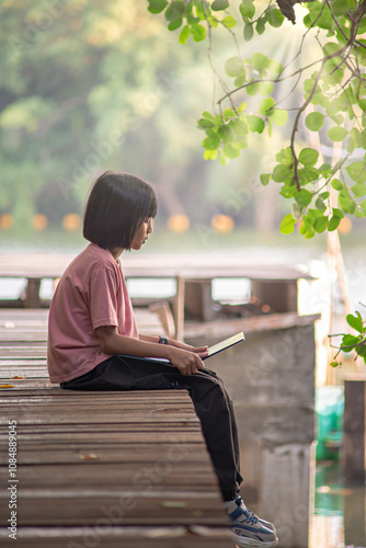 young girl is sitting on a wooden dock by a canal or river in a peaceful and natural setting. She is reading a book as the morning or evening sunlight filters through the green leaves above her head. photo