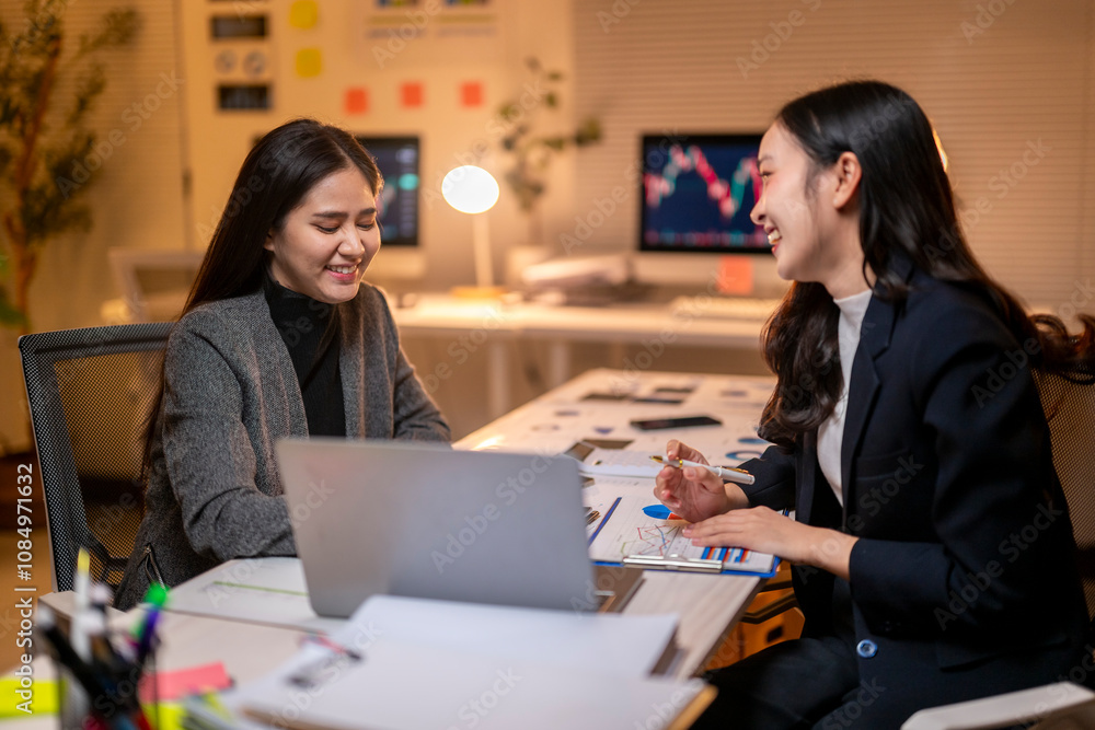 Two women are sitting at a desk, one of them smiling Two women are sitting at a desk, one of them smiling
