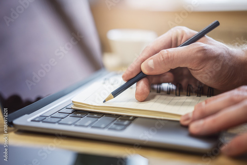 Close-up depiction of a man's hand taking notes in a notepad on a sleek laptop, with a blurred background