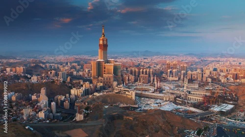 Aerial view of the Abraj Al-Bait Clock Tower and Grand Mosque in Mecca illuminated by the golden light of sunrise, surrounded by the sprawling holy city and mountains