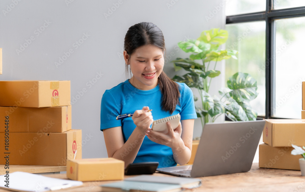 Smiling Woman Working on Laptop: A young woman in a blue shirt sits at a desk in a home office, managing her online business. She's surrounded by cardboard boxes. Smiling Woman Working on Laptop: A young woman in a blue shirt sits at a desk in a home office, managing her online business. She's surrounded by cardboard boxes.