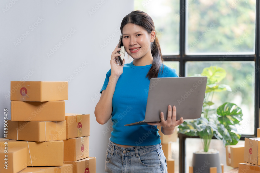 Smiling Businesswoman Managing Orders: A young, Asian entrepreneur confidently manages her online business, smiling while using her laptop and phone amidst a stack of shipping boxes. Smiling Businesswoman Managing Orders: A young, Asian entrepreneur confidently manages her online business, smiling while using her laptop and phone amidst a stack of shipping boxes.