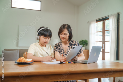 A mother teaches her son to study online at home, using a digital tablet for learning. They sit together at a table with snacks, promoting homeschooling and family bonding.