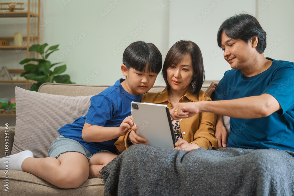 Fototapeta premium Asian parents and children sit together on a sofa at home, laughing and enjoying family time while using various devices like laptops, phones, and tablets, showcasing a tech-savvy lifestyle