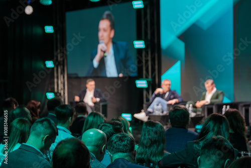 Audience attentively listening to panel discussion at business conference. Blurred stage and vibrant lights create engaging atmosphere for networking and learning.