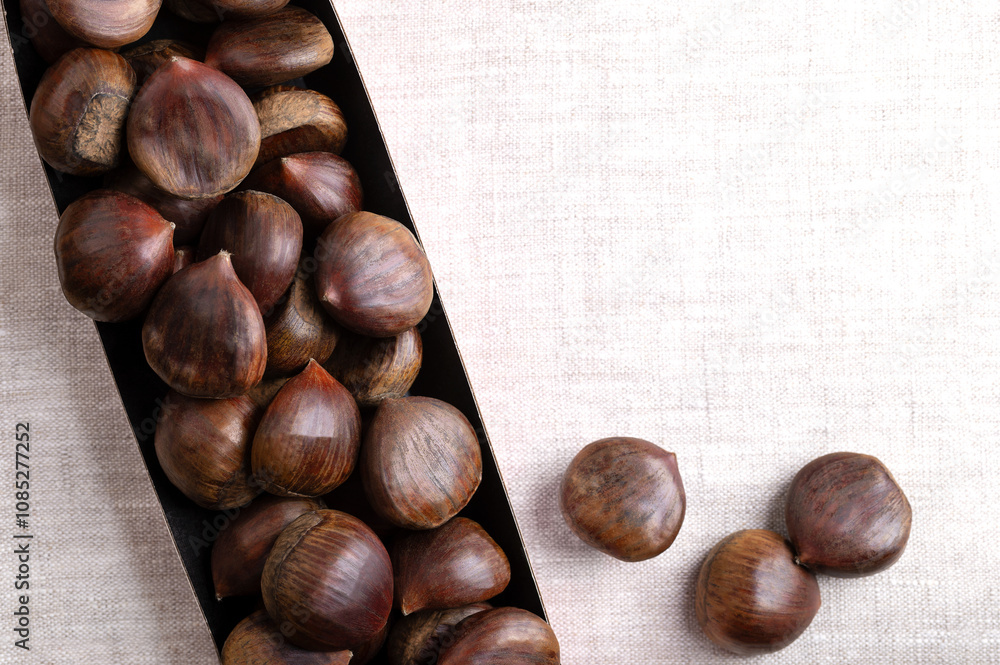 Obraz premium Sweet chestnuts in cardboard punnet on linen, from above, with empty space for text. Castanea sativa, raw, whole and unshelled. Can be eaten raw, candied, cooked, or roasted as warm snack on streets.