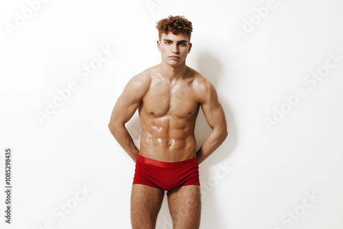 Confident young man posing shirtless in red swim trunks, showcasing fitness and physique. Ideal for health, fitness, and body image concepts.