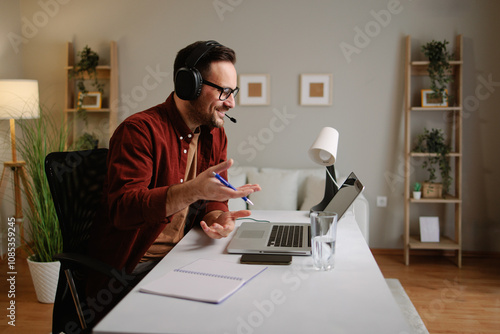 Happy male freelancer talking on headset and taking notes while working remotely from home