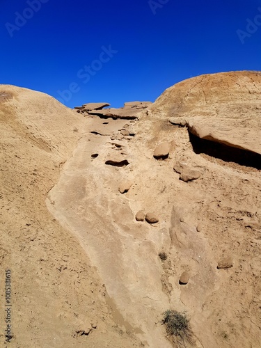 A sandy desert trail flanked by towering rocky formations, bathed in sunlight under the expansive, cloudless blue sky. Perfect for adventurous travelers.