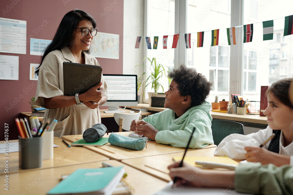 Young smiling teacher with tablet looking at clever schoolboy and answering his question at lesson of English grammar Young smiling teacher with tablet looking at clever schoolboy and answering his question at lesson of English grammar