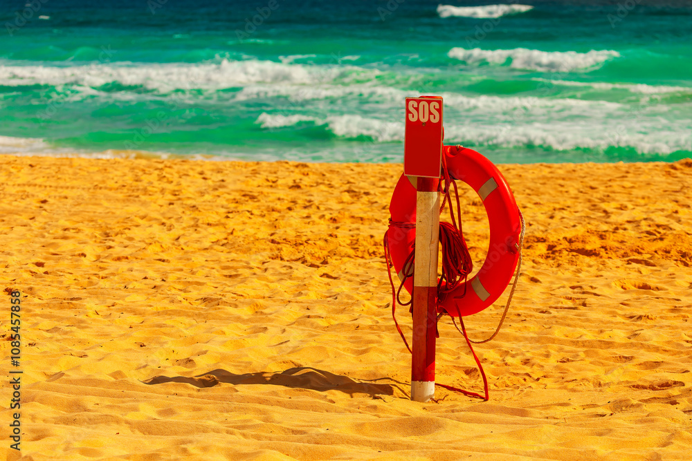 Sandy beach with a lifebuoy attached to wooden post. Inscription SOS provides emergency contact information. Background features the ocean with waves crashing onto shore, indicating a coastal safety