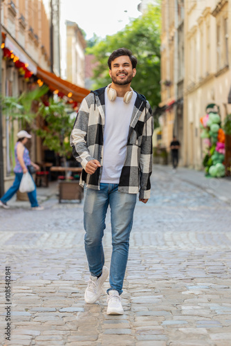 Happy Indian man tourist walking in urban city street background in summer daytime. Young guy traveler smiling having positive good mood enjoying outdoors. Town lifestyles, vacation, holidays trip.