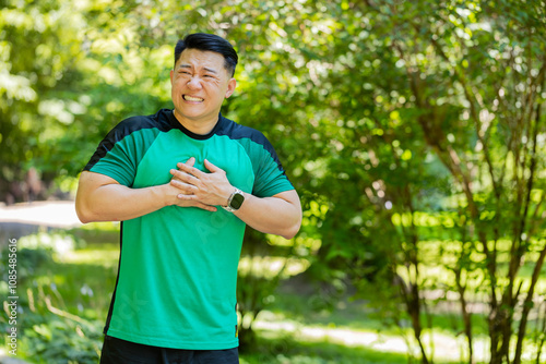 Young adult Asian runner having heart attack or chest pain after running workout in park on sunny summer day. Tired Japanese athlete suffers from tension in the heart. Health care and sports concept.