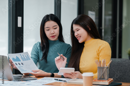 Two Young Women Collaborating in Modern Office Setting with Laptops and Documents, Analyzing Data and Discussing Business Strategies