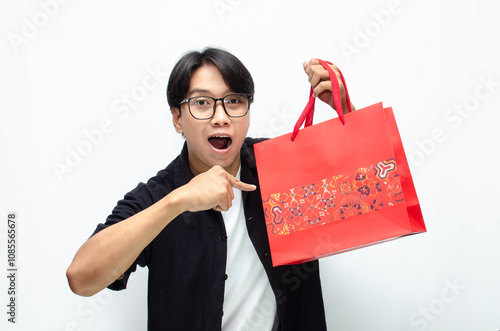 Attractive and stylish young Asian man holding red batik-patterned shopping bags while pointing finger to copy space best for advertisment.