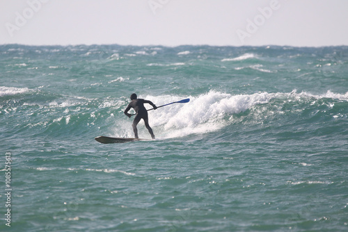 Obraz na płótnie surfer in a wetsuit with a paddle on a board glides along the waves in a strong wind w salonie