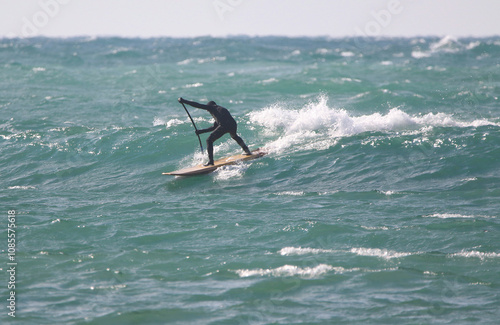 Obraz na płótnie surfer in a wetsuit with a paddle on a board glides along the waves in a strong wind w salonie