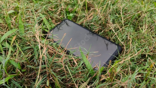 Black smartphone or touchscreen mobile phone with silicone cover protection fall on the park green grass ground floor surface and not being damaged. Closeup slow motion top view.