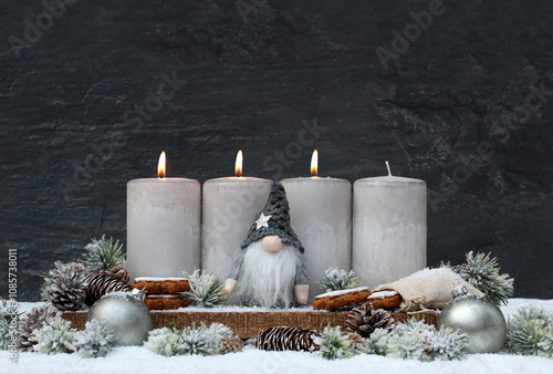 	Dritter Advent: Dekoration mit vier grauen Adventskerzen einem Wichtel und Weihnachtsschmuck im Schnee vor einer dunklen Schieferwand mit Textfreiraum.