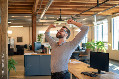 Male employee in a modern office space, performing standing stretches next to his computer, releasing tension.