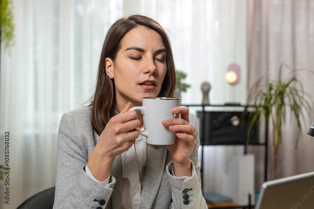 Smiling joyful Caucasian businesswoman drinking a cup of warm hot coffee sitting at workplace home office room in the morning. Happy young girl freelancer enjoying relaxing, taking a break after work. Smiling joyful Caucasian businesswoman drinking a cup of warm hot coffee sitting at workplace home office room in the morning. Happy young girl freelancer enjoying relaxing, taking a break after work.