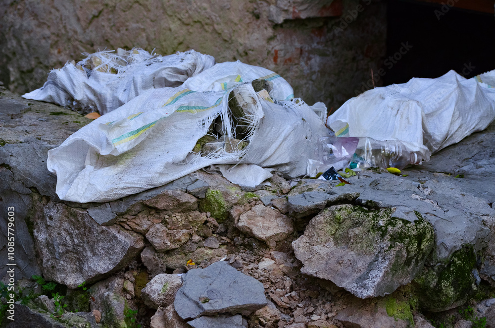A pile of trash is on a rock. The trash includes a white bag with a yellow and green stripe, a plastic bottle, and a glass bottle. The scene is dirty and unkempt. A pile of trash is on a rock. The trash includes a white bag with a yellow and green stripe, a plastic bottle, and a glass bottle. The scene is dirty and unkempt.