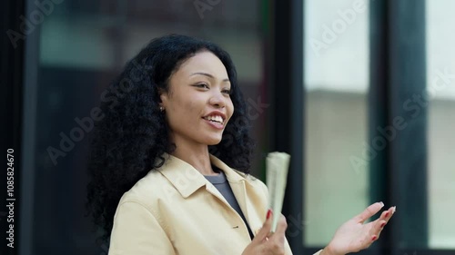 Cheerful woman holding a stack of cash while gesturing with confidence, symbolizing financial success in an urban setting.