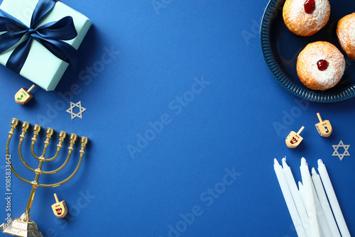 Happy Hanukkah concept. Top view frame of golden menorah, dreidels, doughnuts, candles, and gift box on a blue background. Ideal for festive flat lay style greeting cards or holiday designs photo