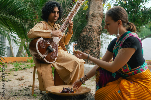 Indian musician in traditional clothes playing sitar festive season outdoor home. Woman in traditional saree hand lighting Diya lamp during Diwali festival. Happy greeting photo. Part of a series