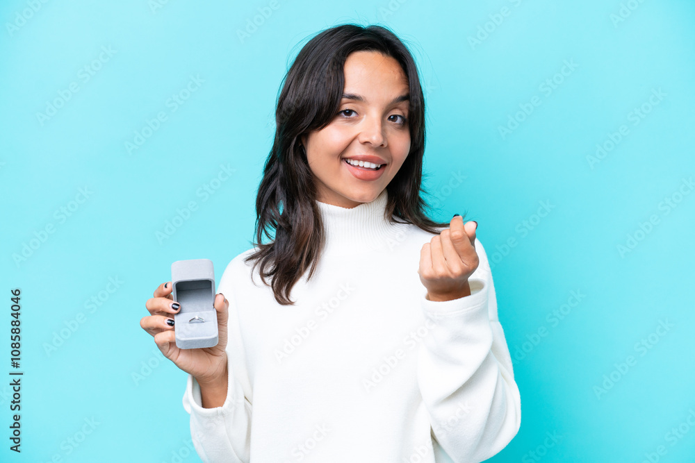 Fototapeta premium Young hispanic woman holding a engagement ring isolated on blue background making money gesture