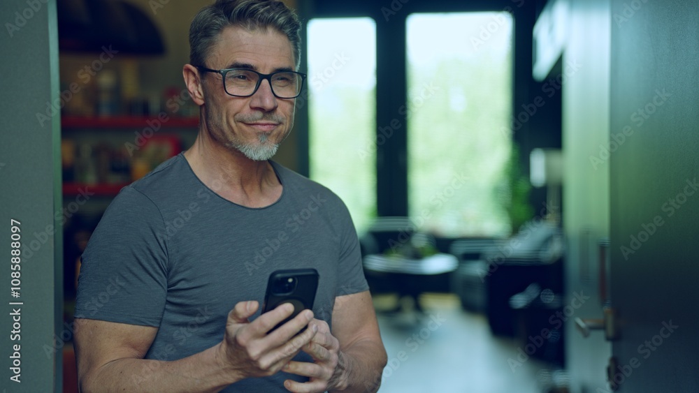 Confident man in 50s standing in modern home interior, wearing casual grey t-shirt, using modern technology with confidence. Portrait of happy mid adult, middle aged male with phone, smiling. Confident man in 50s standing in modern home interior, wearing casual grey t-shirt, using modern technology with confidence. Portrait of happy mid adult, middle aged male with phone, smiling.