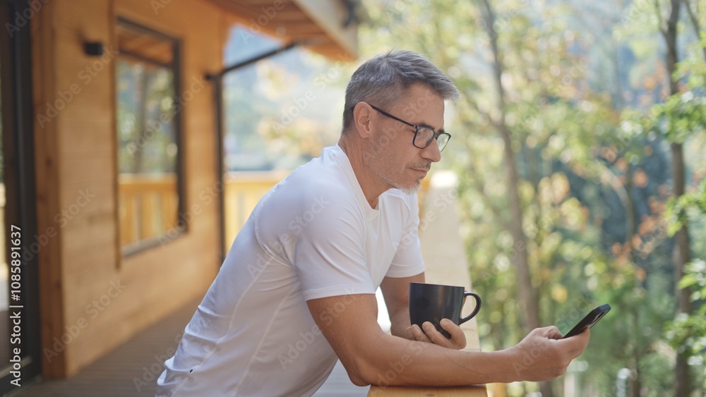 Man in 50s enjoying peaceful morning in wooden cabin. Businessman drinking coffee on balcony of mountain chalet. Entrepreneur working with smartphone. Relaxation and getaway, ideal lifestyle. Man in 50s enjoying peaceful morning in wooden cabin. Businessman drinking coffee on balcony of mountain chalet. Entrepreneur working with smartphone. Relaxation and getaway, ideal lifestyle.